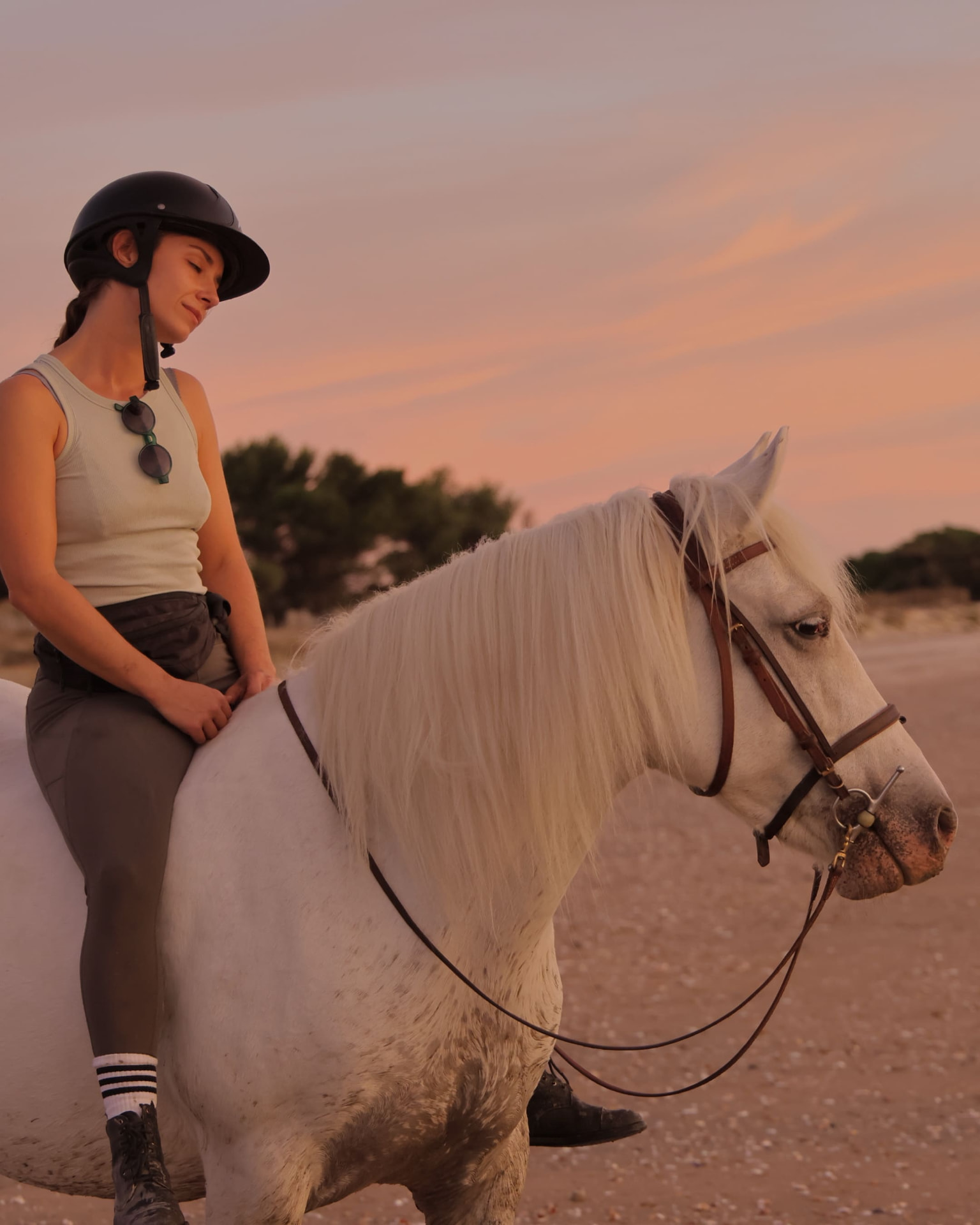 Cavalière casquée sur son cheval blanc, plage au coucher du soleil, ciel rose et orangé